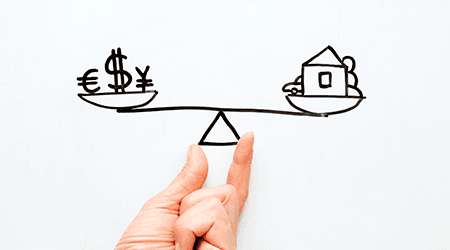 Hand holding a drawing of currency symbols and a house on a weighing scale